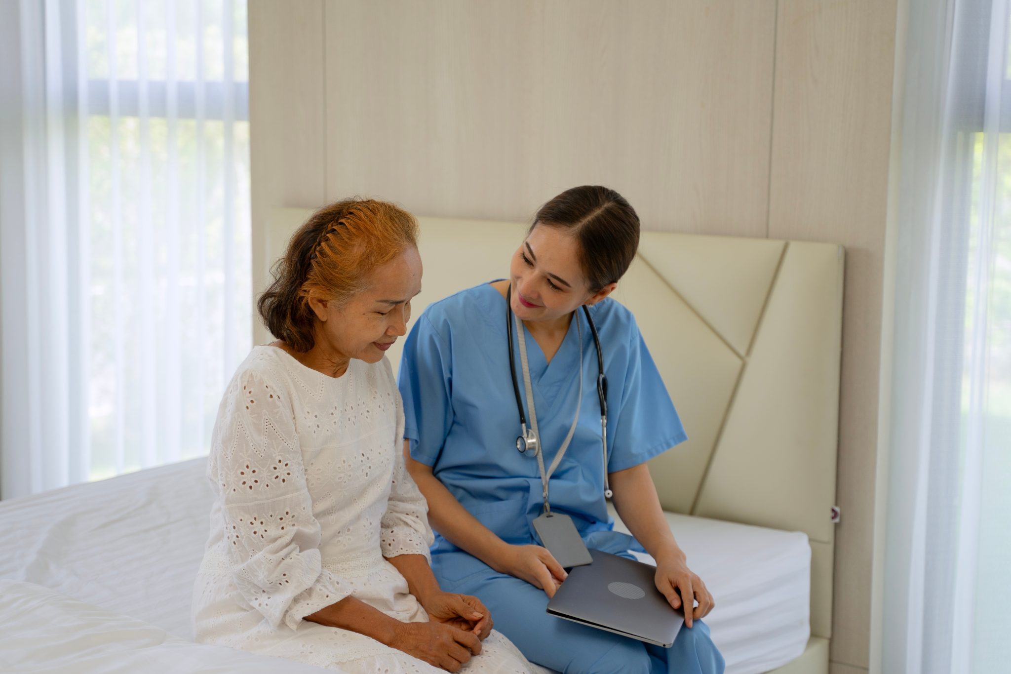 Caregiver assisting an older adult on a bed in an assisted living setting.