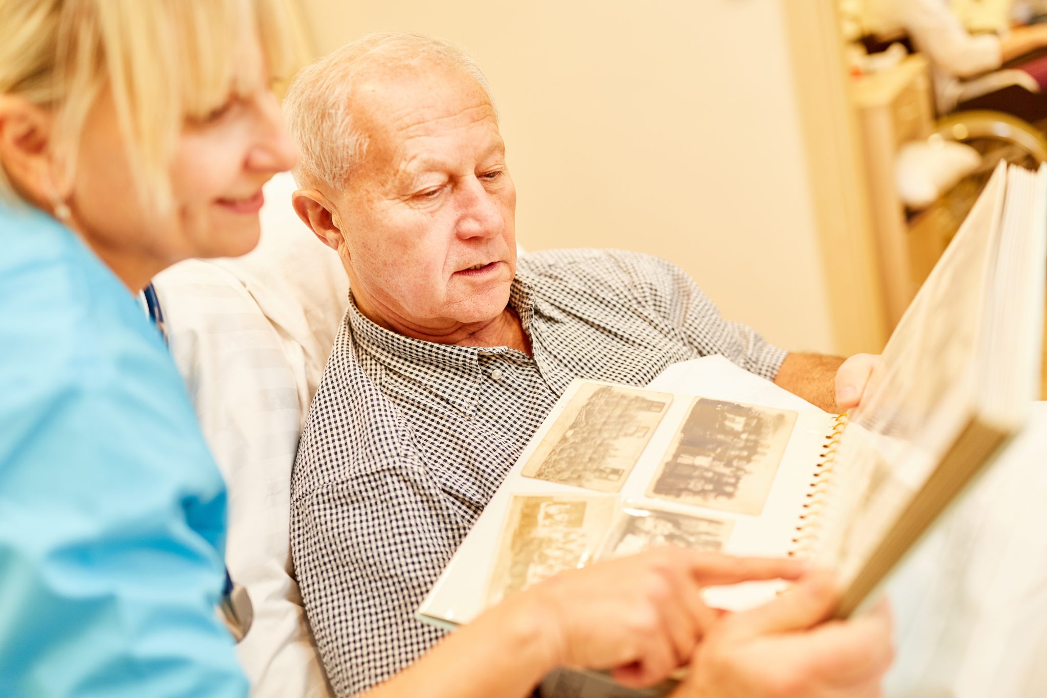 A nursing home staff member in scrubs looks at an open photo album with an older man, both leaning in toward the page
